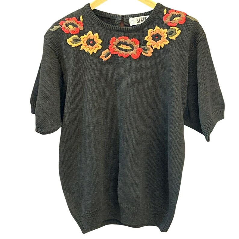 May include: Dark gray short-sleeve sweater with embroidered floral design in red, yellow, and green. The crew neck sweater has a keyhole back and is hanging on a wooden hanger. The word "SEGU" is visible on the back of the sweater.