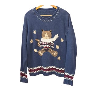 Peut inclure: Un pull en tricot bleu à manches longues orné d'un chat brodé portant un pull à motifs et une écharpe blanche. Le motif comprend des feuilles d'automne brodées, une encolure cousue de blanc et un ourlet à motif géométrique.