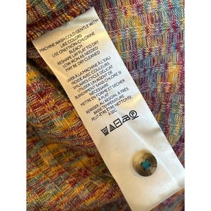 May include: A white care label with instructions for washing and ironing a garment. The label has text in both English and French. The text reads "Machine wash cold gentle with like colors, use only non-chlorine bleach, reshape lay flat to dry, low iron as needed, may be dry cleaned." and "Laver &agrave; la machine &agrave; l'eau froide avec couleurs similaires cycle d&eacute;licat utiliser un agent de blanchiment sans chlore si n&eacute;cessaire mettre en forme et s&eacute;cher &agrave; plat repasser, au besoin, &agrave; tr&egrave;s basse temp&eacute;rature peut-&ecirc;tre &ecirc;tre nettoy&eacute;e &agrave; sec".