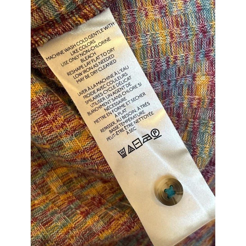 May include: A white care label with instructions for washing and ironing a garment. The label has text in both English and French. The text reads "Machine wash cold gentle with like colors, use only non-chlorine bleach, reshape lay flat to dry, low iron as needed, may be dry cleaned." and "Laver &agrave; la machine &agrave; l'eau froide avec couleurs similaires cycle d&eacute;licat utiliser un agent de blanchiment sans chlore si n&eacute;cessaire mettre en forme et s&eacute;cher &agrave; plat repasser, au besoin, &agrave; tr&egrave;s basse temp&eacute;rature peut-&ecirc;tre &ecirc;tre nettoy&eacute;e &agrave; sec".