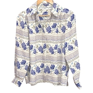 Puede incluir: Camisa de manga larga con estampado floral. La camisa tiene un fondo blanco con flores de hortensia azules y rayas decorativas grises. La camisa está colgada en una percha de madera.