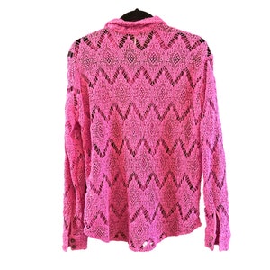 K&ouml;nnte beinhalten: Ein leuchtend pinkfarbenes Langarmhemd mit einem offenen, aufwendigen Spitzendesign. Das Hemd hat einen Kragen und gekn&ouml;pfte Manschetten. Die R&uuml;ckseite des Hemdes ist sichtbar und zeigt das detaillierte Muster. Das Hemd h&auml;ngt an einem schwarzen Kleiderb&uuml;gel.