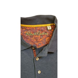 May include: A dark gray polo shirt with a brown and gold collar. The collar has a patterned lining with the text "Robert Graham" in a blue and white font. The shirt has a button closure and a size tag that says "M".