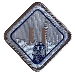 May include: A silver and blue embroidered patch with a diamond shape. The patch features a cityscape with two towers and a blue emblem at the bottom. The border is silver.