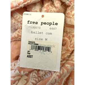 Puede incluir: Una etiqueta blanca con texto negro que dice "free people" y "ballet com" con la talla "M" en una tela de color melocot&oacute;n con un patr&oacute;n floral blanco.
