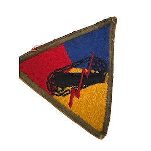 Op de afbeelding: Een driehoekige patch met een rood, blauw en geel ontwerp. De patch heeft een zwarte tank met een rode bliksemschicht en kettingontwerp. De patch is omrand met een groene rand.