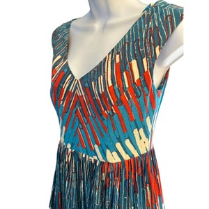 Op de afbeelding: Een mouwloze jurk met een V-hals en een abstracte print in blauw, wit en rood. De jurk heeft een aansluitend bovenlijf en een zwierige rok.