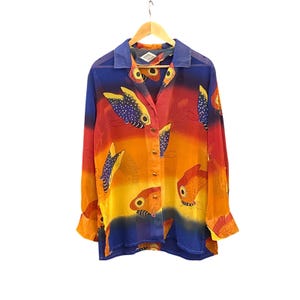 Könnte beinhalten: Ein langärmliges Button-Down-Hemd mit einem lebendigen Fischmuster. Das Hemd hat einen Farbverlauf aus Blau, Orange und Gelb. Die Fische sind orange mit schwarzen und weißen Details. Das Hemd hängt an einem Holzbügel.