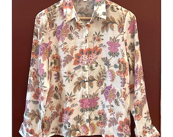 Puede incluir: Camisa abotonada con estampado floral en tonos rosa, naranja, morado y verde sobre fondo crema. La camisa de manga larga está colgada de una percha de madera.