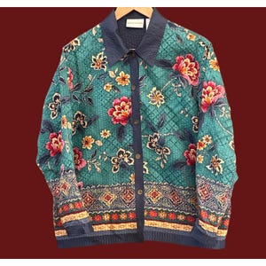 May include: A teal quilted jacket with a floral pattern featuring red, pink, and yellow flowers. The jacket has a navy blue collar and trim, with a button-up front. The floral design is set against a diamond-patterned background.