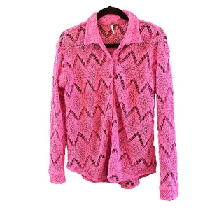 K&ouml;nnte beinhalten: Ein leuchtend pinkfarbenes Langarmhemd mit Knopfleiste und Kragen. Das Hemd besteht aus einem zarten, offen gewebten Spitzenstoff mit einem detaillierten Blumen- und geometrischen Muster. Das Hemd h&auml;ngt an einem schwarzen Kleiderb&uuml;gel.
