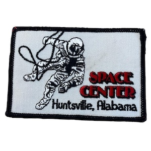 May include: A rectangular embroidered patch with a white background and a black border. It features a black-stitched astronaut figure with a tether, the red text "SPACE CENTER," and the black text "Huntsville, Alabama" printed below it.