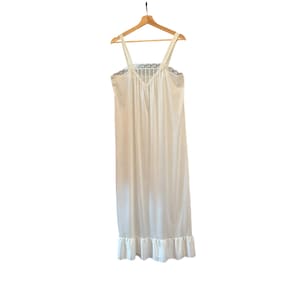 May include: A floor-length white nightgown featuring a V-neckline with delicate lace trim along the bodice and shoulder straps. The garment has a smooth, flowing silhouette and a ruffled hemline at the bottom, hanging from a wooden hanger.