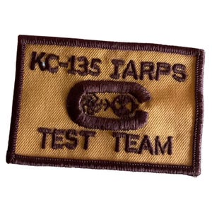 May include: A rectangular patch with a gold background and a dark brown border. The patch features the text "KC-135 TARPS TEST TEAM" in dark brown, along with a stylized emblem.