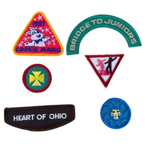 May include: A collection of six embroidered patches including a triangular 1987 Cookie Magic patch, an arched Bridge to Juniors patch, a circular yellow cross patch, a triangular patch with figures, a brown Heart of Ohio patch, and a blue circular patch.