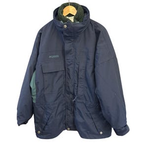 May include: A navy blue Columbia winter jacket with a high collar and a green fleece lining. The jacket has a front pocket and the Columbia logo on the chest. The jacket is hanging on a wooden hanger.