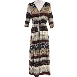 Op de afbeelding: Een lange, gelaagde maxi-jurk met een kleurrijk, geprint ontwerp. De jurk heeft een V-hals, 3/4 mouwen en een knoopsluiting aan de voorkant. De stof is licht en soepel.