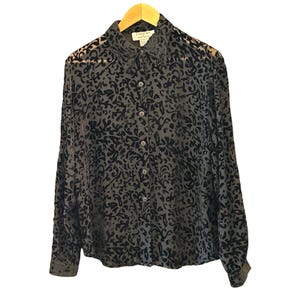 May include: A long-sleeve button-down shirt in a sheer black fabric featuring a dark, raised velvet-like floral pattern throughout. The garment has a standard collar, a front button placket with matching buttons, and a label reading Essentials.