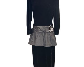 May include: A black velvet dress with long sleeves and a grey peplum skirt. The dress has an oval-shaped embellishment with pearl accents at the waist. The dress is a vintage style.