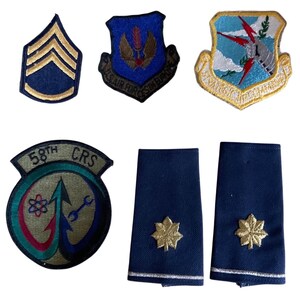 May include: A collection of military patches and insignia, including a sergeant's stripes, several embroidered emblems with various designs, and two dark blue shoulder boards with gold-colored starburst details. The patches feature text and detailed imagery.
