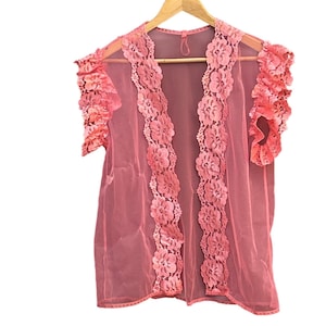Könnte beinhalten: Ein transparentes, korallenfarbenes Kleid mit floraler Spitzenborte und Rüschenärmeln hängt an einem Holzbügel. Das offene Design hat eine zarte, feminine Ästhetik. Die Spitzenborte ist in einem dunkleren Korallton gehalten und setzt einen Kontrast zum Kleidungsstück.