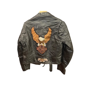 May include: Black leather motorcycle jacket with an embroidered eagle patch on the back. The patch features a brown eagle with outstretched wings and the Harley-Davidson Cycles logo. The jacket has a classic biker style with a notched collar.
