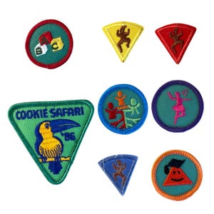 Pode incluir: Uma coleção de oito emblemas de tecido bordado em várias formas e cores. Os designs incluem blocos de letras, figuras a dançar, um tucano com o texto COOKIE SAFARI 86, uma bailarina e um chapéu de graduação sobre um triângulo.