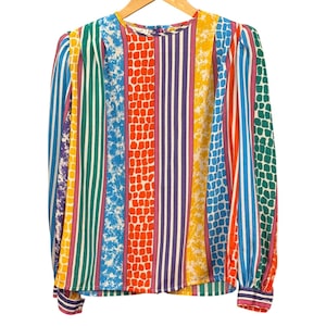 Könnte beinhalten: Eine langärmelige Bluse mit einem lebendigen vertikalen Muster aus mehrfarbigen Streifen und strukturierten Blöcken. Das Design umfasst abwechselnde Bänder in Grün, Blau, Gelb, Orange und Violett, wobei einige Abschnitte ein gesprenkeltes oder gitterartiges Muster aufweisen.