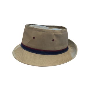 May include: A tan bucket hat featuring a flat crown and a wide brim. The hat is accented with a horizontal navy blue and maroon striped ribbon band encircling the base of the crown. Two small, dark-colored ventilation eyelets are visible on the side.