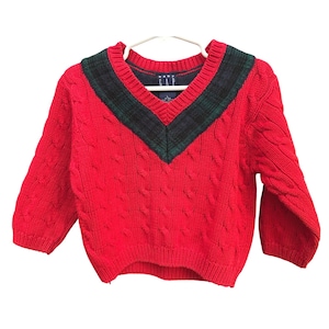 Puede incluir: Un suéter rojo de punto trenzado con cuello en V con un diseño de cuadros verdes y negros. El suéter tiene mangas largas y se muestra en una percha blanca. La etiqueta de la marca GAP es visible.
