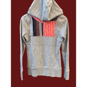 Peut inclure: Un sweat &agrave; capuche gris avec une fermeture &eacute;clair compl&egrave;te et une capuche. Le dos du sweat-shirt pr&eacute;sente un patch d&eacute;coratif avec des rayures verticales dans les tons de rouge, rose, noir et bleu. Le sweat-shirt a des manches longues et des poignets c&ocirc;tel&eacute;s.