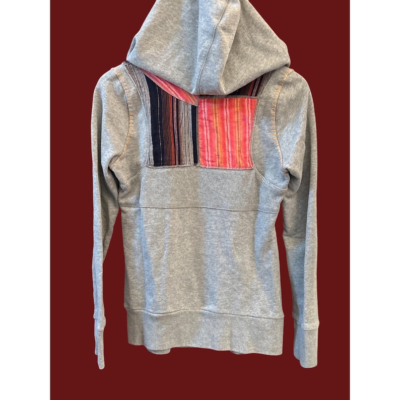Peut inclure: Un sweat &agrave; capuche gris avec une fermeture &eacute;clair compl&egrave;te et une capuche. Le dos du sweat-shirt pr&eacute;sente un patch d&eacute;coratif avec des rayures verticales dans les tons de rouge, rose, noir et bleu. Le sweat-shirt a des manches longues et des poignets c&ocirc;tel&eacute;s.