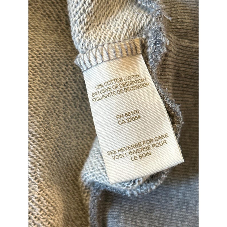 Peut inclure: Gros plan sur un tissu gris avec une &eacute;tiquette blanche attach&eacute;e. L'&eacute;tiquette indique "100% COTTON/COTON EXCLUSIVE OF DECORATION/ EXCLUSIVIT&Eacute; DE D&Eacute;CORATION" et comprend les num&eacute;ros RN et CA. Le texte "SEE REVERSE FOR CARE VOIR L'INVERSE POUR LE SOIN" est &eacute;galement visible.