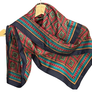 May include: A patterned silk scarf in shades of red, teal, and navy blue. The scarf features a repeating geometric design with a navy blue border. The scarf is draped over a wooden hanger.