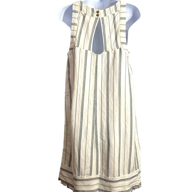 Puede incluir: Vestido blanco sin mangas con finas rayas negras y un escote en la espalda en forma de ojo de cerradura. El vestido tiene un dobladillo festoneado.