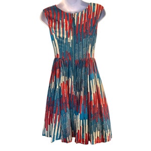 Op de afbeelding: Een mouwloze jurk met een abstract patroon in blauw, rood en wit. De jurk heeft een aansluitend bovenlijf en een volle rok.