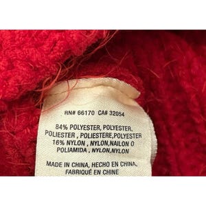 May include: Close-up of a red knitted fabric with a white tag. The tag displays text including "RN# 66170 CA# 32054" and material composition: 84% polyester and 16% nylon. The tag also indicates the item was made in China.