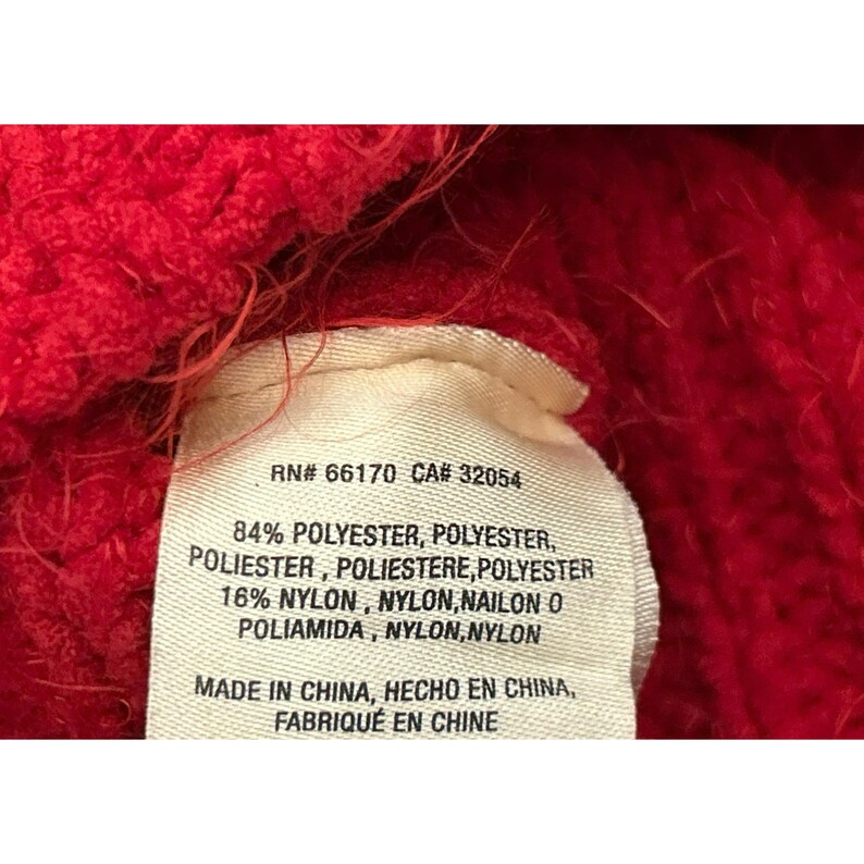 May include: Close-up of a red knitted fabric with a white tag. The tag displays text including "RN# 66170 CA# 32054" and material composition: 84% polyester and 16% nylon. The tag also indicates the item was made in China.