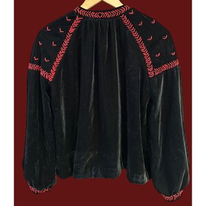 May include: Black velvet blouse with long sleeves and red embroidered detailing. The embroidery is featured around the neckline and shoulders. The blouse is hanging on a wooden hanger.