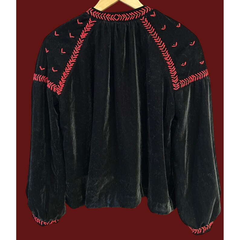 May include: Black velvet blouse with long sleeves and red embroidered detailing. The embroidery is featured around the neckline and shoulders. The blouse is hanging on a wooden hanger.