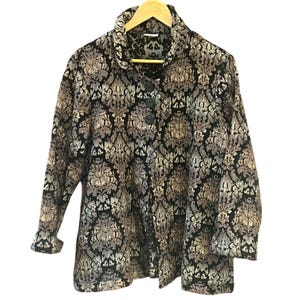 May include: A long-sleeved jacket with a black, gray, and tan floral pattern. The jacket has a large collar, three buttons, and is hanging on a wooden hanger. The fabric has a textured appearance.