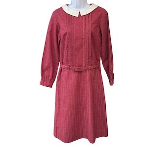 May include: A long-sleeved red dress featuring a white Peter Pan collar, vertical pinstripes, and a matching fabric belt at the waist. The bodice includes vertical pleats, and the sleeves are finished with buttoned cuffs.