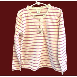 Puede incluir: Camisa de manga larga a rayas blancas y rosas, colgada de una percha blanca. La camisa tiene un cuello con volantes y una tapeta con botones. El nombre de la marca "crewcuts" es visible en la etiqueta.