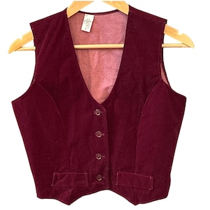 May include: A deep burgundy velvet vest featuring a V-neckline, four matching buttons down the center front, and two rectangular welt pockets at the waist. The garment is displayed on a wooden hanger and includes a white interior clothing tag.