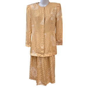 May include: A two-piece set consisting of a long-sleeved jacket and a matching skirt in a light tan color. The fabric features a repeating geometric pattern and white leaf-like motifs. The jacket has a round neckline and a front button closure.