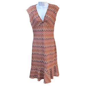 May include: A sleeveless, knee-length dress with a V-neck and a repeating zigzag pattern in shades of orange, pink, and brown. The dress has a fitted bodice and a slightly flared skirt, creating a flattering silhouette.