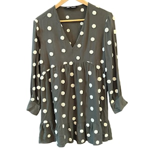 May include: A dark olive green dress with white polka dots. The dress has a V-neck, long sleeves with cuffs, and a ruffled hem. The dress is hanging on a wooden hanger.