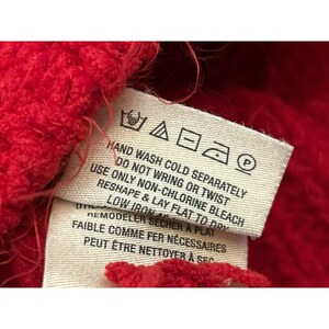 May include: Close-up of a red knitted fabric with a care label attached. The label provides washing instructions, including hand wash cold, do not wring or twist, and reshape and lay flat to dry. The label also includes ironing instructions.