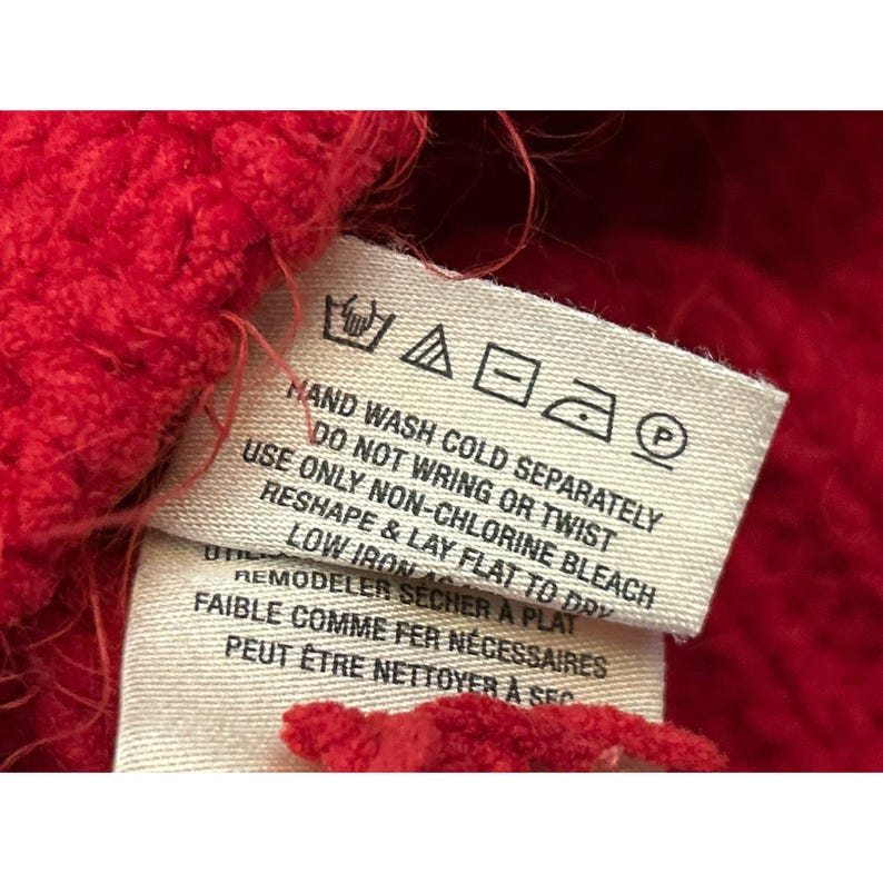 May include: Close-up of a red knitted fabric with a care label attached. The label provides washing instructions, including hand wash cold, do not wring or twist, and reshape and lay flat to dry. The label also includes ironing instructions.