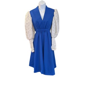 May include: A royal blue dress with a V-neck and a fitted waist. The dress features white, sheer, long sleeves with a floral pattern. The sleeves have a slight puff at the shoulder and are gathered at the wrist. The dress is displayed on a white mannequin.